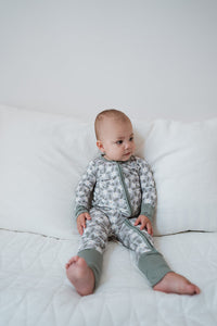 A baby sitting on a white quilted bed wearing a long-sleeve zip-up pajama set with a gray and white animal print and sage green cuffs and hem.