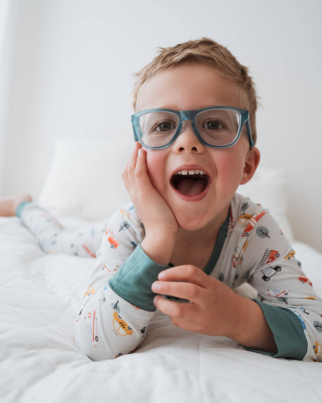 A young boy with short blond hair and blue eyeglasses lies on a white bed, smiling with his mouth open. He is wearing a light gray long-sleeve pajama shirt with a colorful cartoon print and teal cuffs.