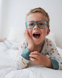 A young boy with short blond hair and blue eyeglasses lies on a white bed, smiling with his mouth open. He is wearing a light gray long-sleeve pajama shirt with a colorful cartoon print and teal cuffs.
