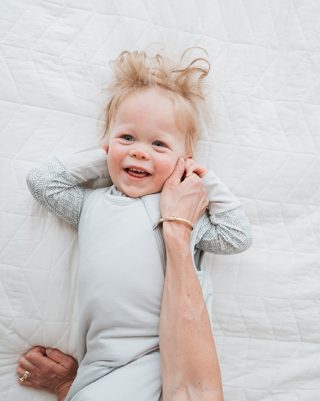 A baby wearing a light gray long-sleeve romper lies on a white quilted bed while an adult's hand gently holds the baby's head. The baby is also wearing a gray knit blanket underneath.