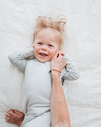 A baby wearing a light gray long-sleeve romper lies on a white quilted bed while an adult's hand gently holds the baby's head. The baby is also wearing a gray knit blanket underneath.