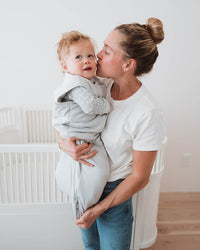 A woman in a white t-shirt and blue jeans holds a baby wearing a light gray wrap carrier in a bright, modern nursery with white cribs and light wood floors.