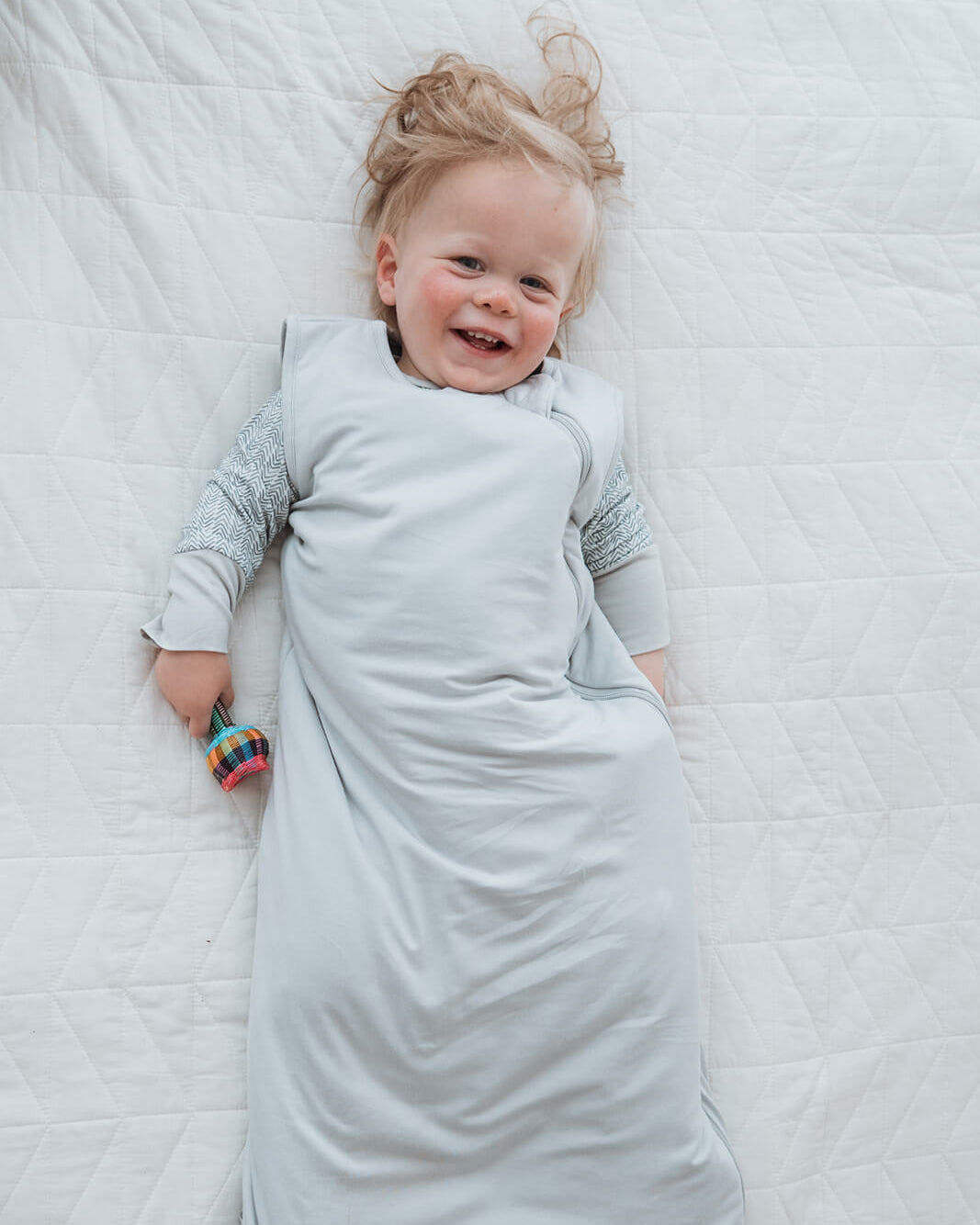 A smiling baby lying on a white quilted blanket, wrapped in a light gray sleep sack over a long-sleeve knit top.