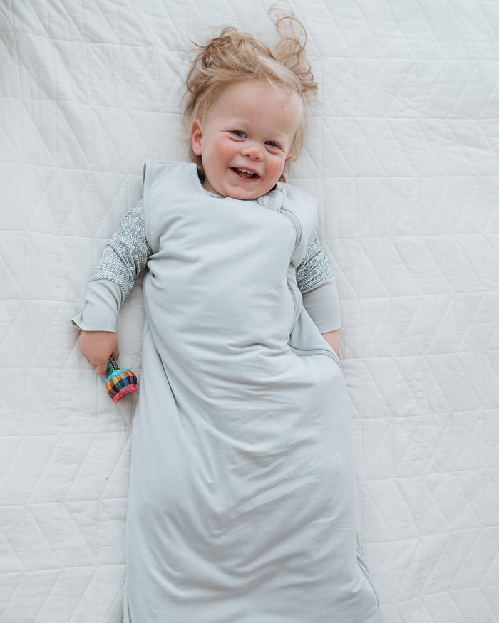 A smiling baby lying on a white quilted blanket, wrapped in a light gray sleep sack over a long-sleeve knit top.