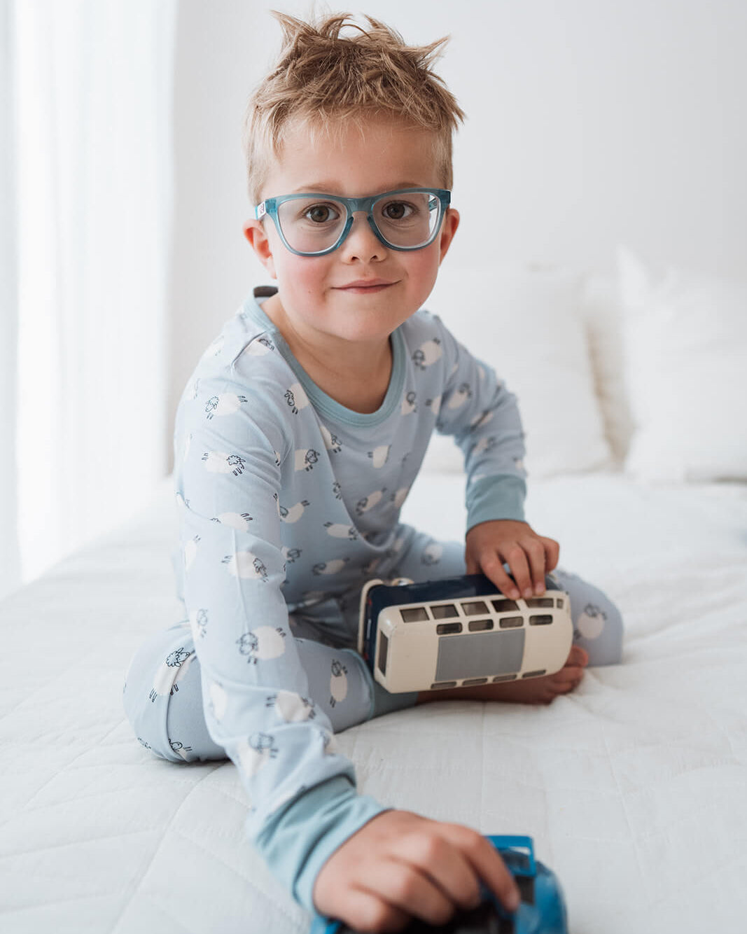 A young boy with short blond hair and blue glasses sits on a white bed, wearing light blue pajamas with a small cloud pattern and holding a toy radio.