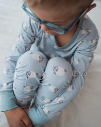A child wearing light blue long-sleeve pajamas with a white sheep pattern, sitting on a white blanket and looking down while wearing translucent blue glasses.