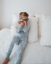 A young boy wearing light blue patterned pajamas and blue glasses lies on a white bed, holding a white plush toy and wiping his nose with a tissue.