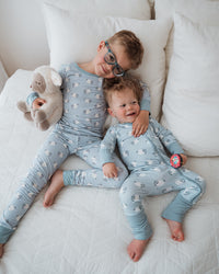 Two young children lying on a white quilted bed wearing matching light blue pajamas with a small white animal print. The older child wears glasses and holds a beige stuffed animal, while the younger child holds a small pink toy.