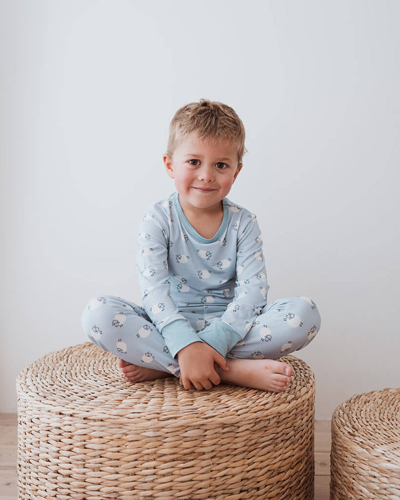 A young child sitting cross-legged on a round woven ottoman in a bright room, wearing a light blue long-sleeve pajama top and matching pajama pants with a small white pattern.