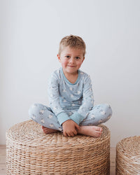 A young child sitting cross-legged on a round woven ottoman in a bright room, wearing a light blue long-sleeve pajama top and matching pajama pants with a small white pattern.
