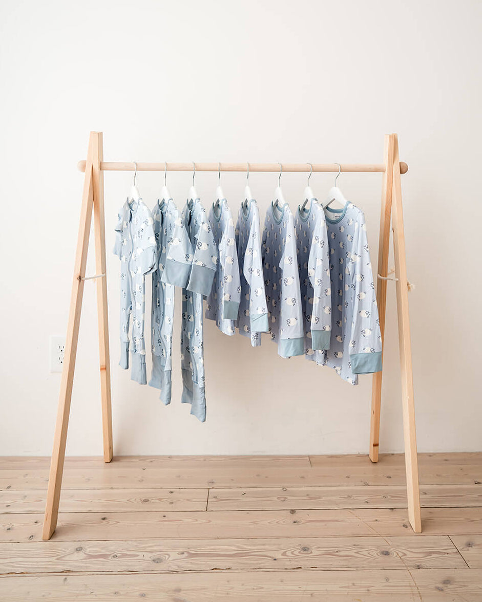 A set of light blue baby garments with white patterns hanging on a minimalist wooden A-frame rack against a plain off-white wall and light wood floor.
