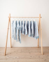 A set of light blue baby garments with white patterns hanging on a minimalist wooden A-frame rack against a plain off-white wall and light wood floor.