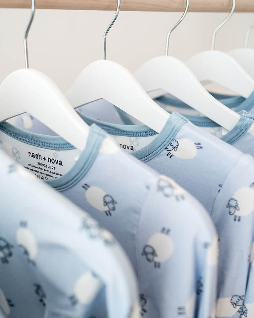 A row of light blue baby long-sleeve shirts with a white sheep pattern, hanging on white hangers from a wooden clothing rack.