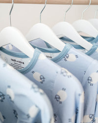 A row of light blue baby long-sleeve shirts with a white sheep pattern, hanging on white hangers from a wooden clothing rack.