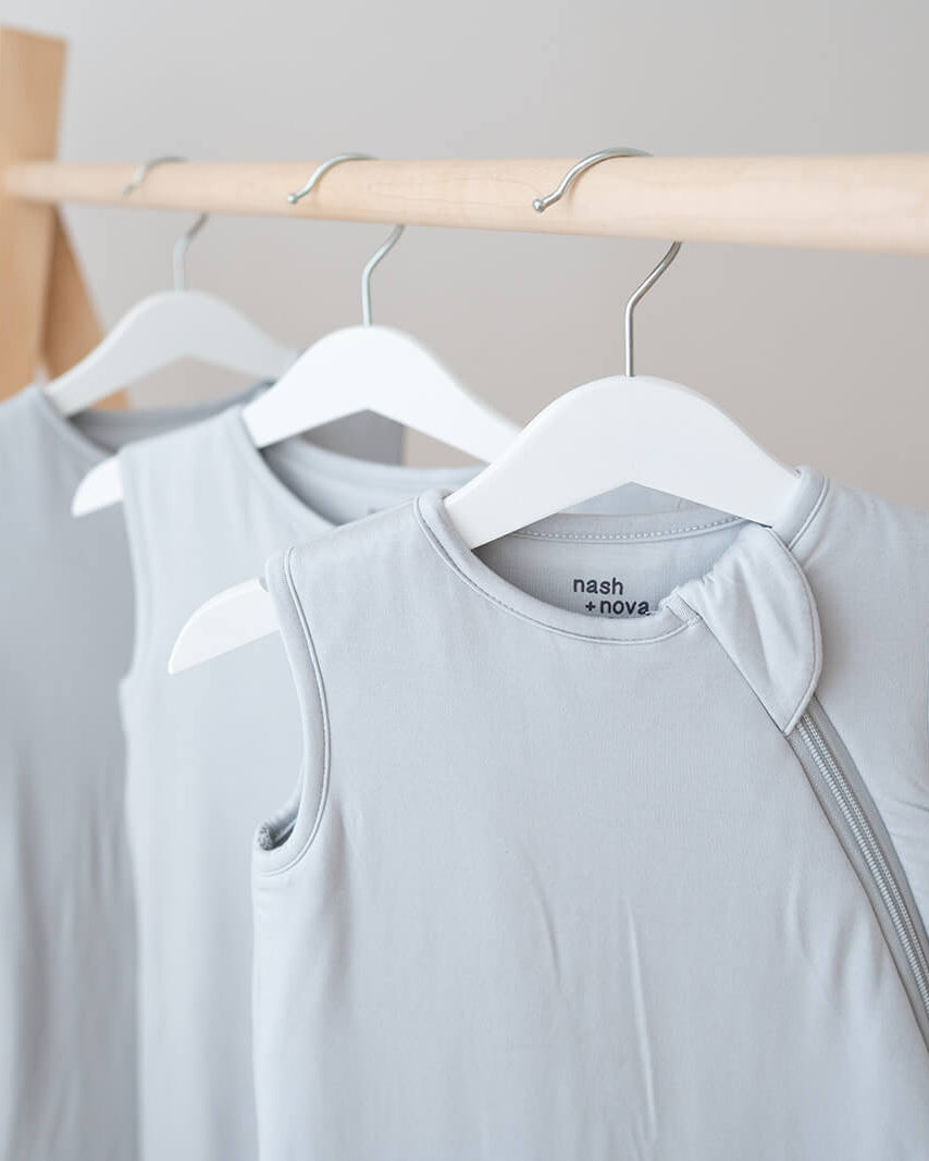 Two light gray sleeveless tank tops hanging on white hangers from a wooden clothing rack against a soft neutral background.