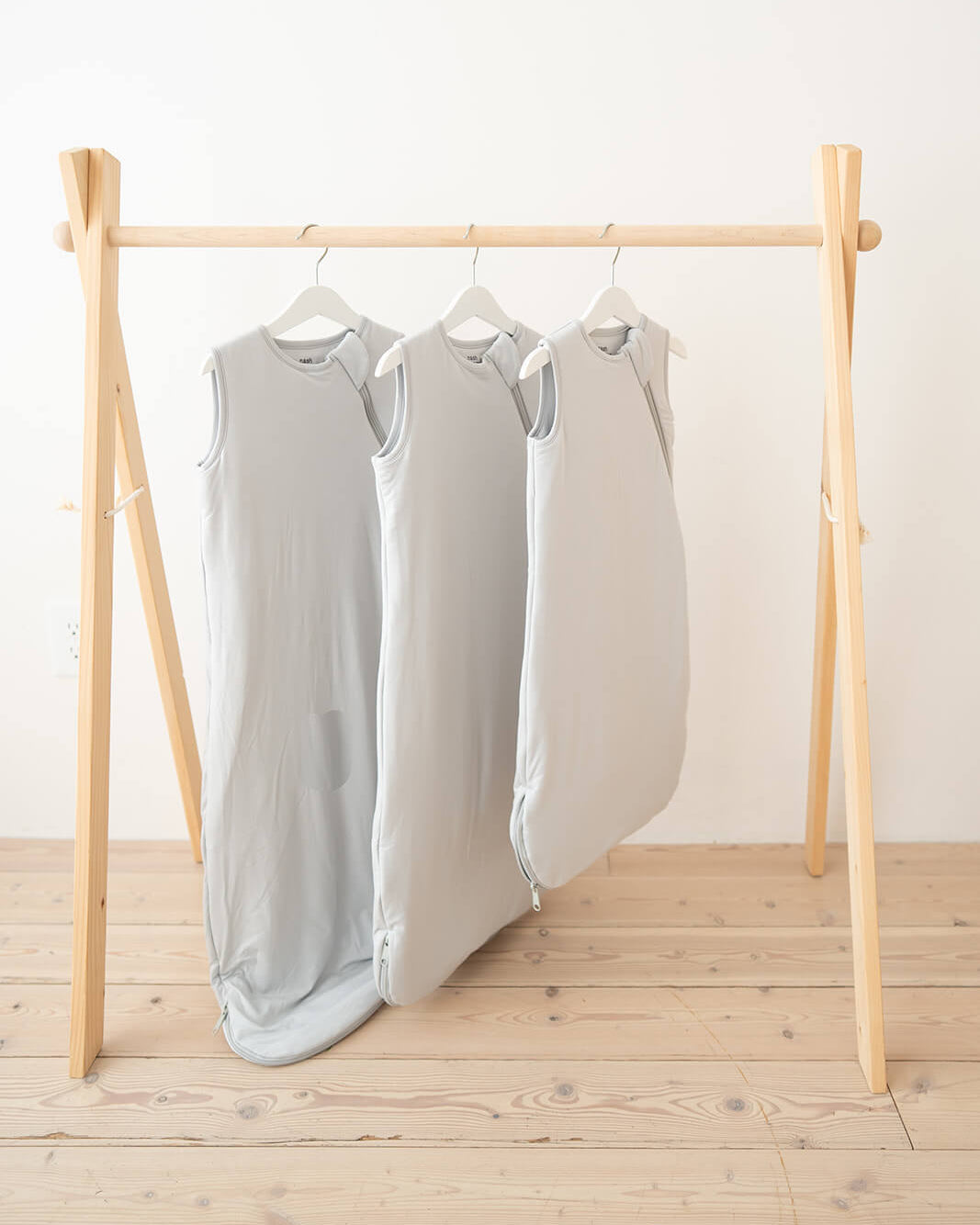 Three light gray sleeveless sleep sacks hanging on a wooden A-frame clothing rack over a light wood floor against a white wall.