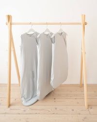 Three light gray sleeveless sleep sacks hanging on a wooden A-frame clothing rack over a light wood floor against a white wall.