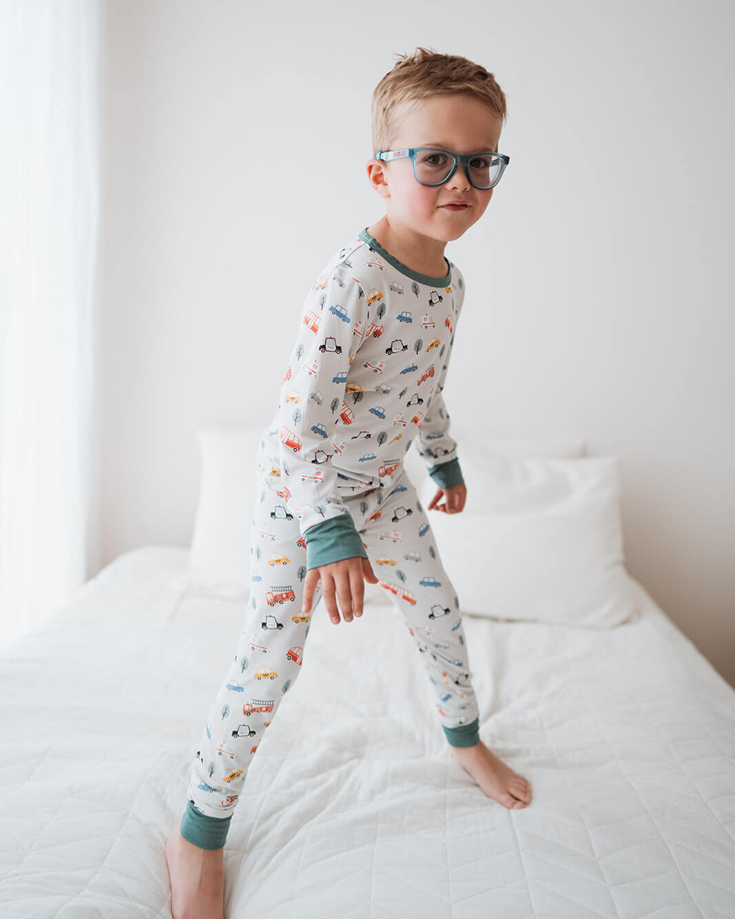 A young boy wearing a long-sleeve pajama set with a colorful car pattern, standing on a white bed in a bright bedroom.