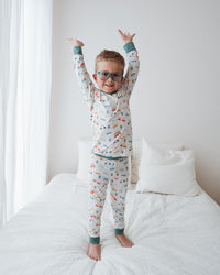 Child in pajamas standing on a bed with arms raised in a bedroom setting