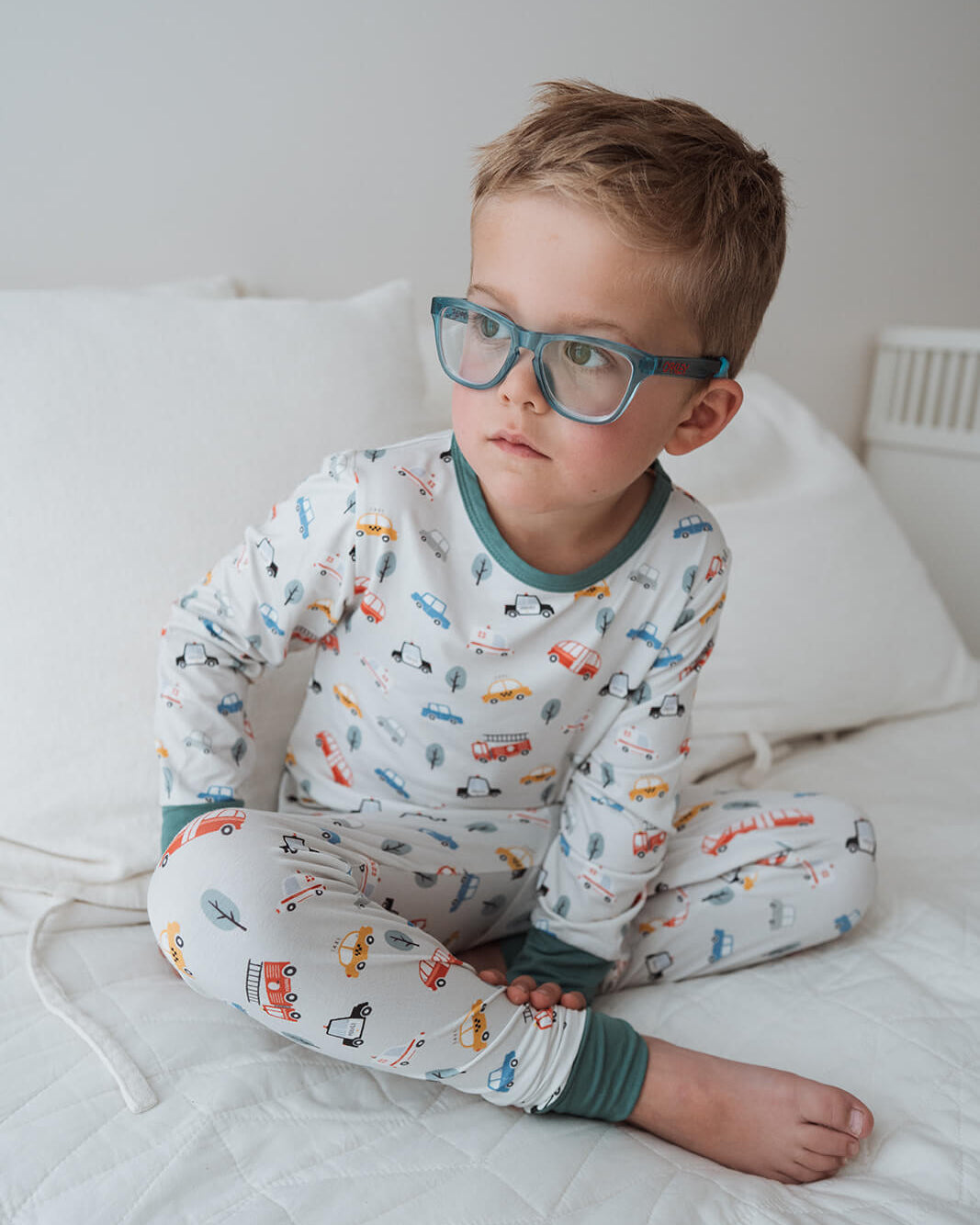 A young boy with short brown hair and blue-framed glasses sits cross-legged on a white quilted bed. He is wearing a long-sleeve pajama set with a white base and a colorful print of small vehicles, with sage green trim at the collar, cuffs, and hem.
