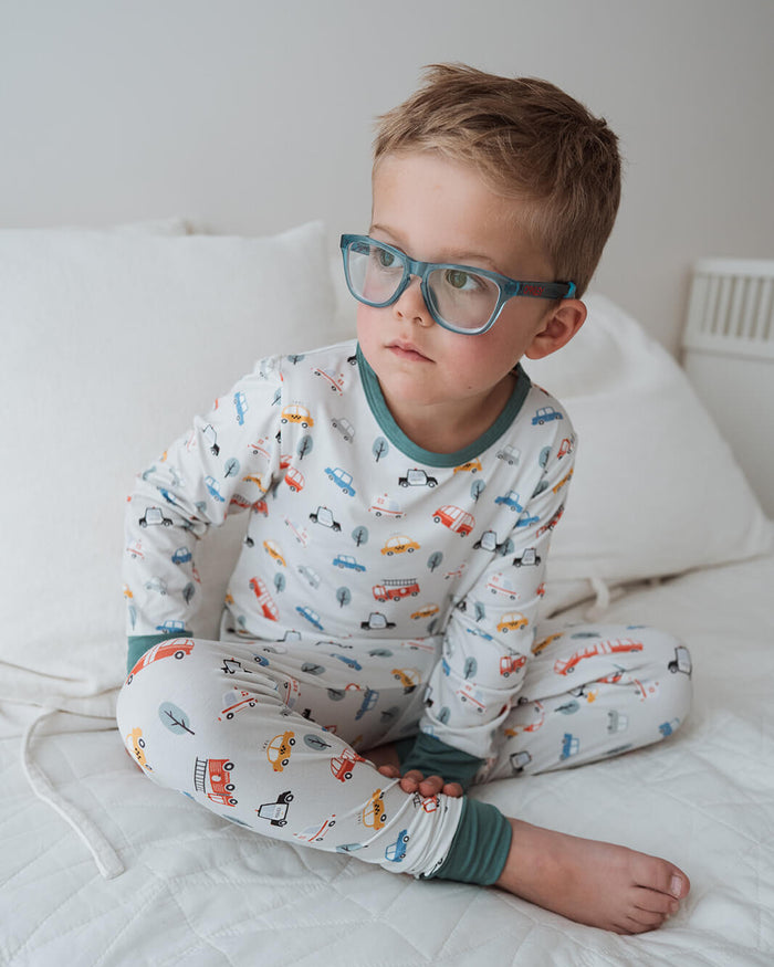 A young boy with short brown hair and blue-framed glasses sits cross-legged on a white quilted bed. He is wearing a long-sleeve pajama set with a white base and a colorful print of small vehicles, with sage green trim at the collar, cuffs, and hem.