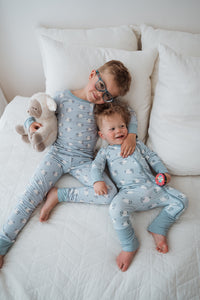 Two young children lying on a white quilted bed wearing matching light blue pajamas with a small white animal print. The older child wears glasses and holds a beige stuffed animal, while the younger child holds a small pink toy.