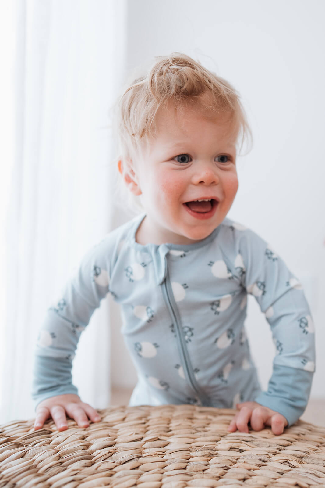A smiling toddler with blonde hair wearing a light blue long-sleeve zip-up onesie with a white sheep pattern, crawling on a woven straw mat.