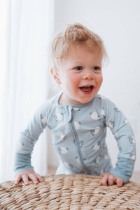 A smiling toddler with blonde hair wearing a light blue long-sleeve zip-up onesie with a white sheep pattern, crawling on a woven straw mat.
