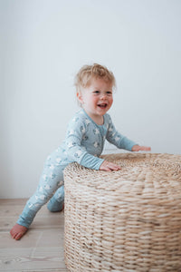 A toddler wearing light blue long-sleeve pajamas with a white dalmatian print is smiling while leaning on a large woven wicker ottoman in a bright room with a white wall and light wood floor.