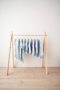 A set of light blue patterned baby garments hanging on a minimalist wooden A-frame clothing rack against a plain off-white wall and light wood floor.