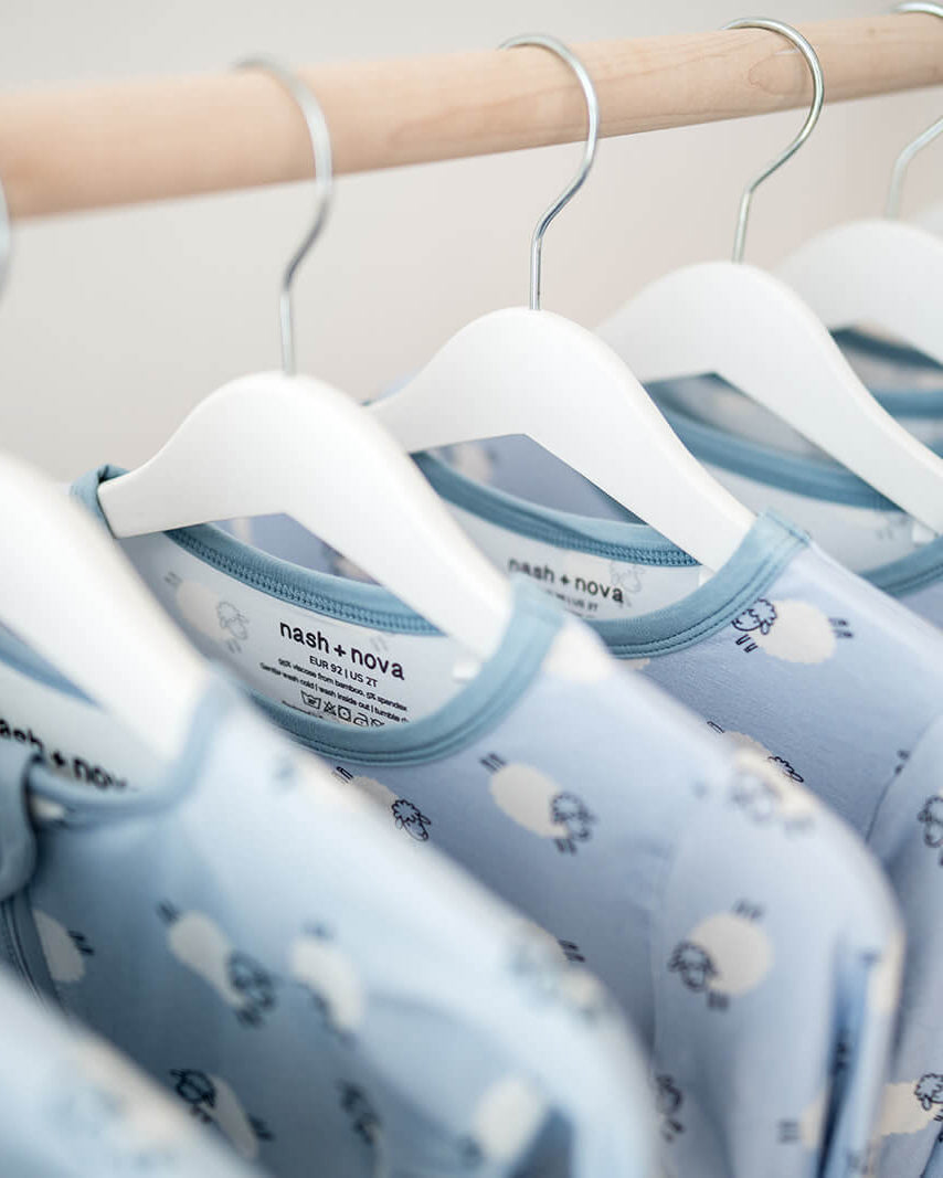 A close-up of several light blue baby long-sleeve shirts with a white cloud pattern hanging on white hangers from a wooden clothing rack.