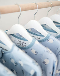 A close-up of several light blue baby long-sleeve shirts with a white cloud pattern hanging on white hangers from a wooden clothing rack.