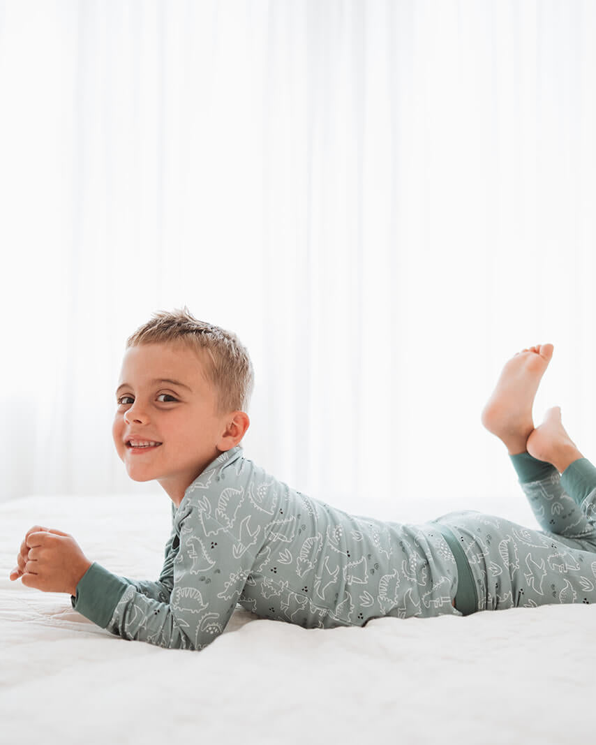 A young boy smiling while lying on a white bed, wearing a long-sleeve pajama set in a muted sage green with a subtle pattern.