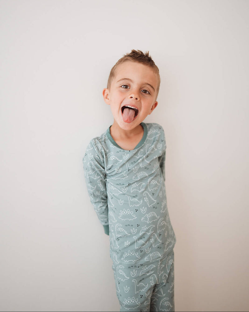 A young boy wearing a light blue-green long-sleeve pajama set with a white dinosaur pattern, sticking his tongue out against a plain off-white background.