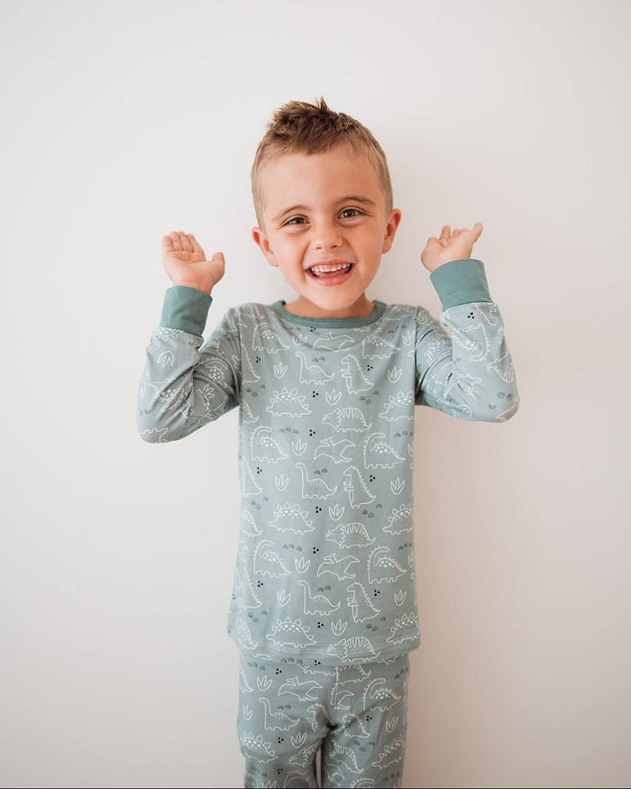 A young boy smiling and posing with his hands raised, wearing a light sage green long-sleeve pajama set with a white dinosaur pattern against a plain white background.