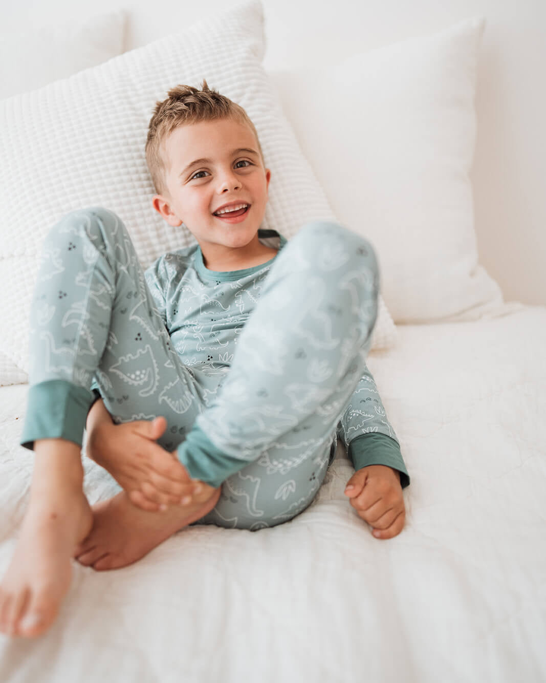 A young boy smiling while sitting on a white bed, wearing a light blue pajama set with a subtle dinosaur pattern.