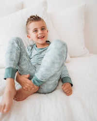 A young boy smiling while sitting on a white bed, wearing a light blue pajama set with a subtle dinosaur pattern.