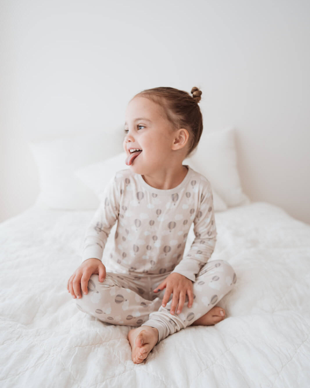 Child wearing a patterned onesie sitting on a white bed.