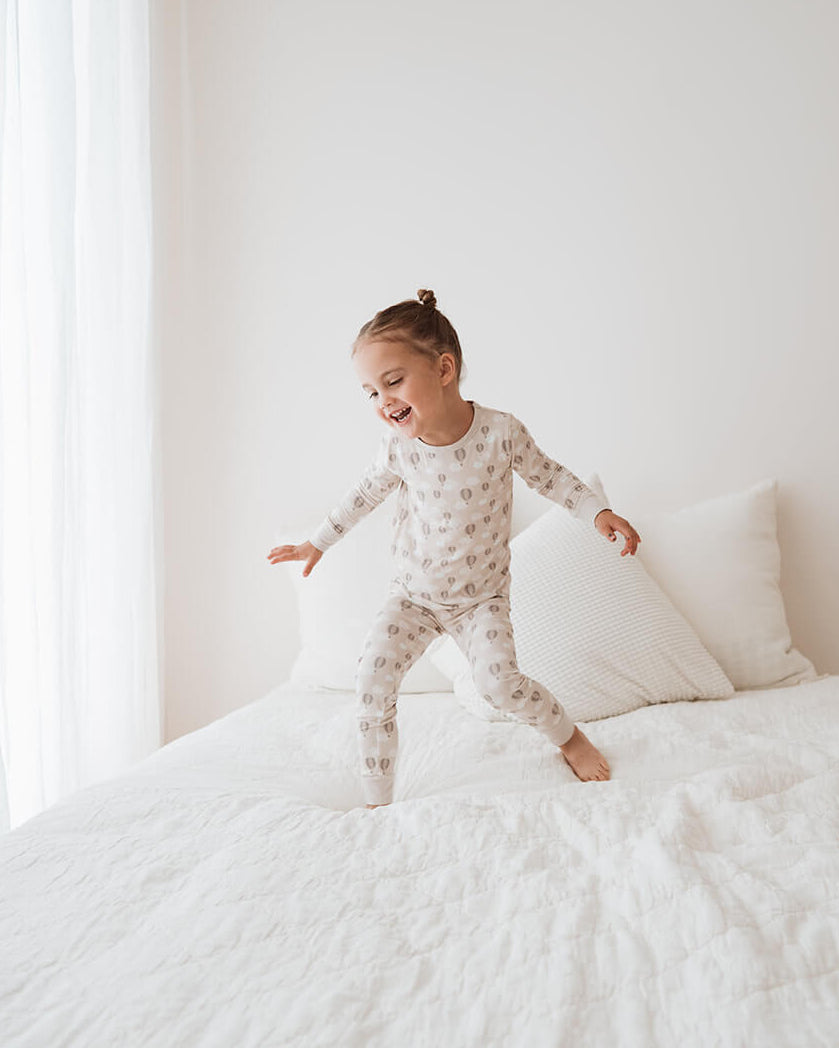 A toddler girl wearing a light beige and white patterned long-sleeve pajama set jumps on a white bed in a bright, minimalist bedroom.