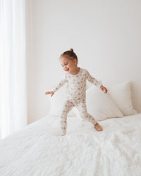A toddler girl wearing a light beige and white patterned long-sleeve pajama set jumps on a white bed in a bright, minimalist bedroom.