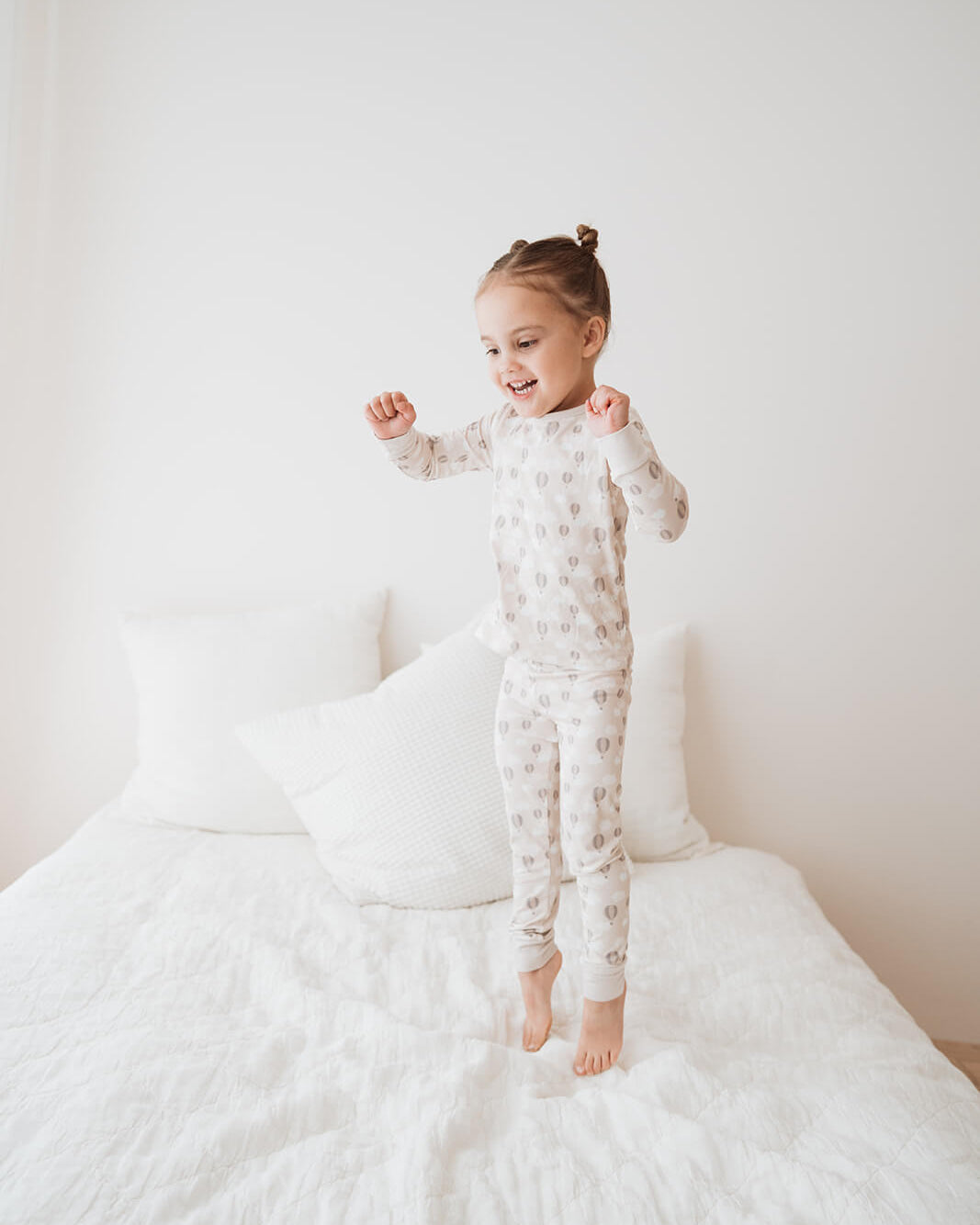 A young child stands barefoot on a white bed, wearing a light beige and white patterned long-sleeve pajama set. The background is a clean, minimalist white bedroom.