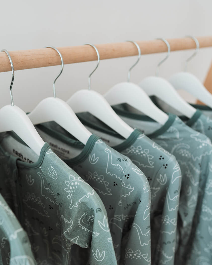 A row of sage green patterned baby t-shirts hanging on white hangers from a light wood clothing rack.