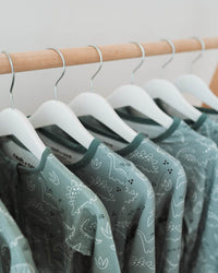 A row of sage green patterned baby t-shirts hanging on white hangers from a light wood clothing rack.