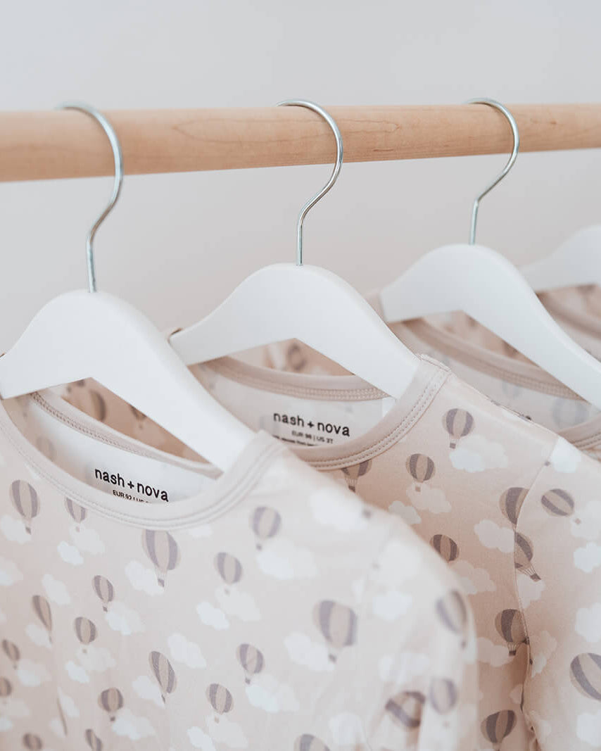A close-up of several long-sleeve baby tops hanging on white hangers from a light wood clothing rack. The tops are a warm beige with a soft pattern of gray and white abstract shapes.