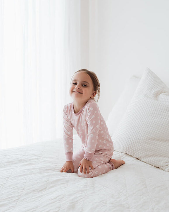 A young child wearing a light pink long-sleeve pajama set sits on a white bed with white pillows in a bright, minimalist bedroom.