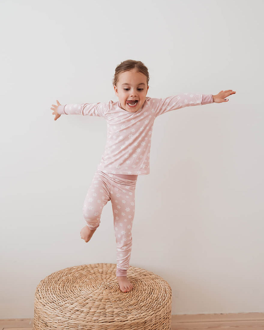 A young girl with short brown hair stands barefoot on a round woven ottoman, balancing with outstretched arms. She is wearing a light pink long-sleeve pajama top and matching pink patterned pajama pants against a plain white wall and light wood floor.