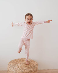 A young girl with short brown hair stands barefoot on a round woven ottoman, balancing with outstretched arms. She is wearing a light pink long-sleeve pajama top and matching pink patterned pajama pants against a plain white wall and light wood floor.