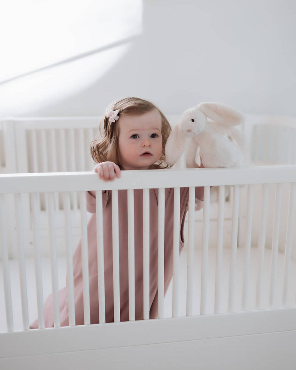 A toddler girl in a dusty pink long-sleeve romper stands inside a white wooden crib, holding onto the white railing next to a white plush bunny toy.