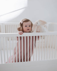 A toddler girl in a dusty pink long-sleeve romper stands inside a white wooden crib, holding onto the white railing next to a white plush bunny toy.
