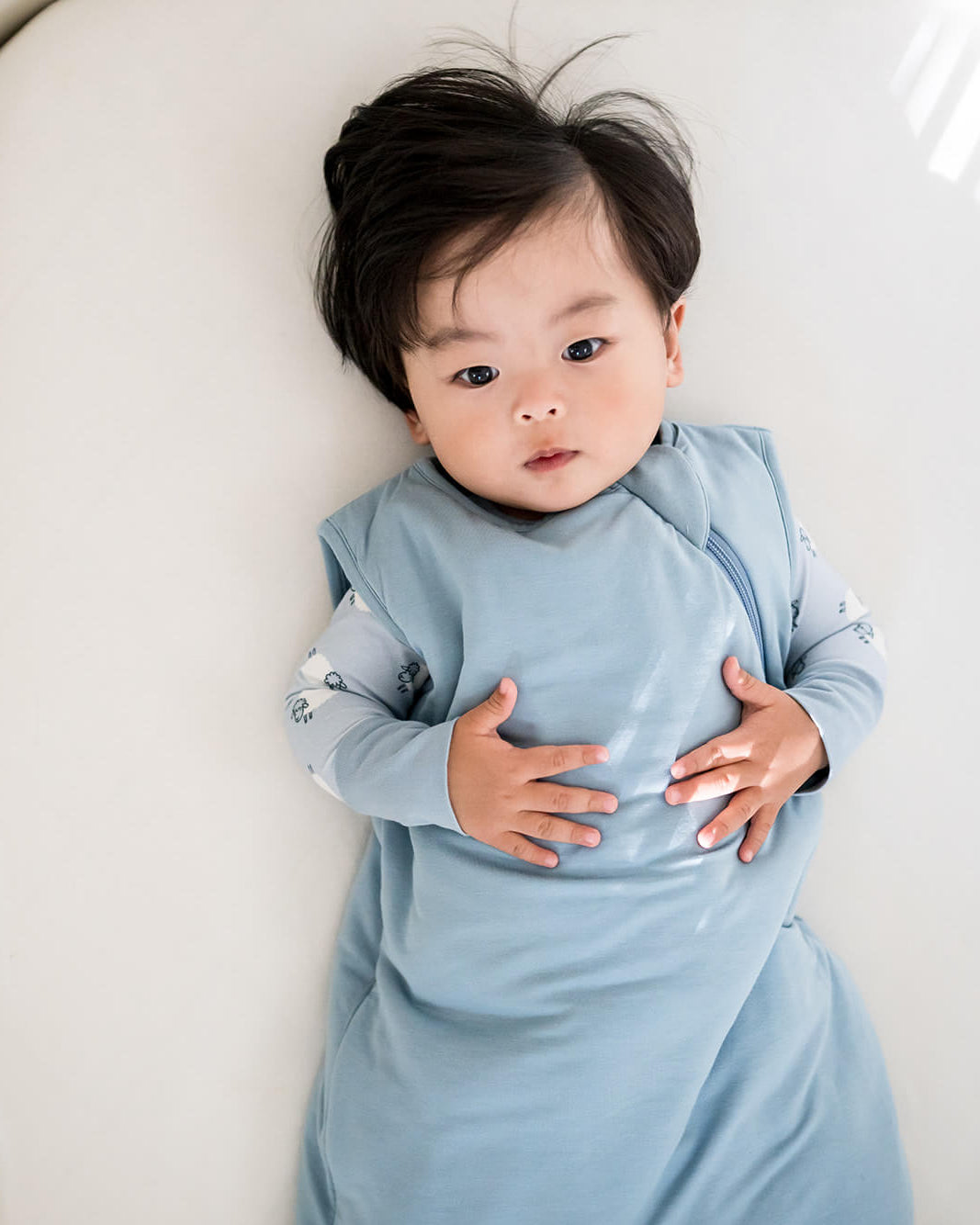 A baby lying on a white changing pad wearing a light blue long-sleeve sleep sack.